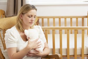 A sad looking woman sitting next to an empty cot cuddling a bear